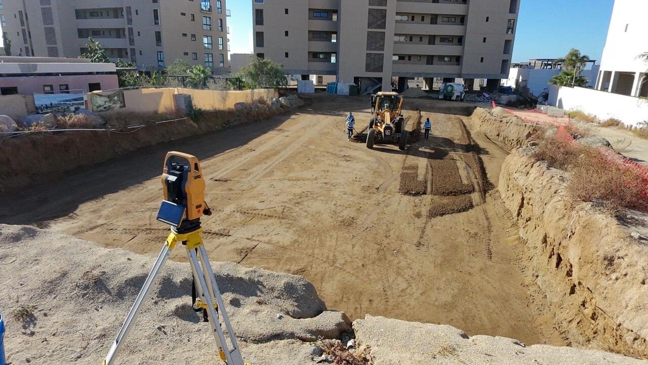 Estación total junto a un frente de terracerías y edificios en construcción
